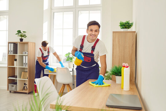 Janitors working in office premises. Two professional cleaners from commercial cleaning service sanitize all surfaces in office. Happy smiling men in workwear wiping wooden tables and shelves together