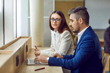 Fototapeta premium Two people meet and communicate in office. Young woman and man in suits sitting together at table by window, using laptop computer, working on business project and talking about ways to improve it