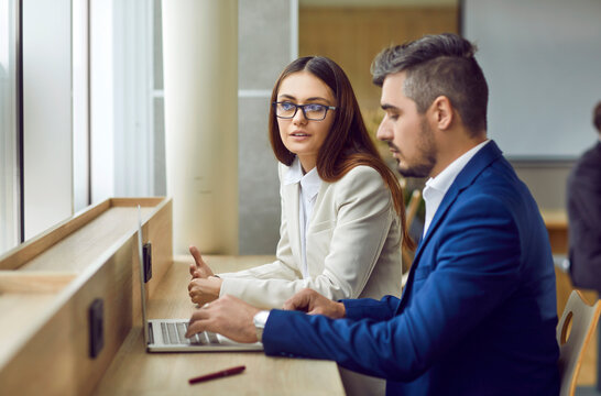 Two People Meet And Communicate In Office. Young Woman And Man In Suits Sitting Together At Table By Window, Using Laptop Computer, Working On Business Project And Talking About Ways To Improve It