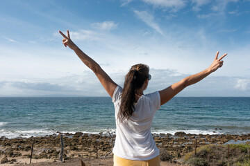 Authentic woman expressing confidence and waving victory signs with open arms under the beautiful sky and sea.