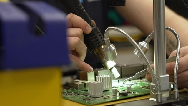 A young man in a mustard shirt and cap works in dim light at a table and solders wires with a soldering iron. Working with a soldering iron from which smoke comes, engineering activities.
