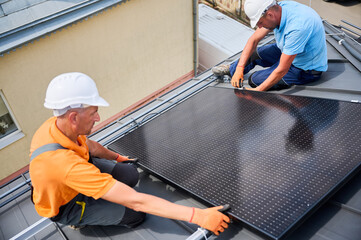 Workers building solar panel system on rooftop of house. Two men installers in helmets installing...