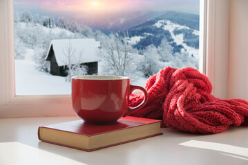 Cozy winter still life: mug of hot tea and opened book with warm plaid on modern windowsill against...