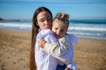Young mon gently hugs her little kid girl, standing with her eyes closed on the sandy beach against...