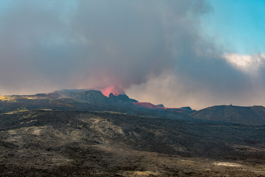 View across a hardened black grey lava field with lava and smoke erupting from a volcano in the distance - Iceland, Geldingadalir near Grindavik - june 2021