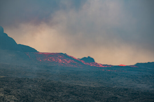 View across a hardened black grey lava field with lava and smoke erupting from a volcano in the distance - Iceland, Geldingadalir near Grindavik - june 2021