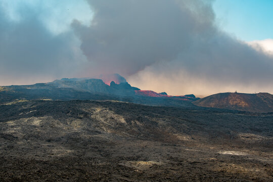 View across a hardened black grey lava field with lava and smoke erupting from a volcano in the distance - Iceland, Geldingadalir near Grindavik - june 2021
