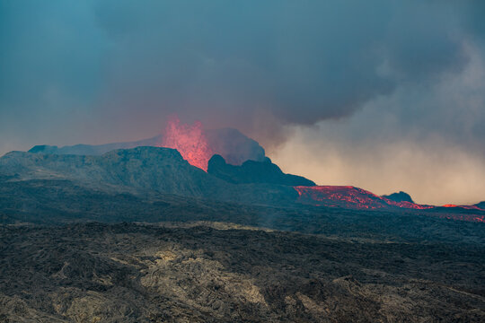 View across a hardened black grey lava field with lava and smoke erupting from a volcano in the distance - Iceland, Geldingadalir near Grindavik - june 2021