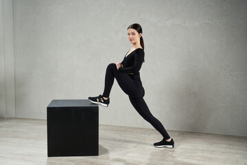 Slim dark-haired woman exercising on black wooden cube in fitness studio. Side view of fit girl...