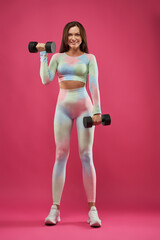 Smiling Caucasian middle-aged fitness woman lifting dumbbell weights. Front view of beautiful slim lady with dumbbells in hands in front of hot pink background. Concept of sport, lifestyle, health.