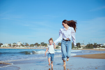 Caucasian happy young mother and daughter smiling while running on the beach, leaving footsteps on the wet sand