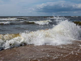 Close-up shot of big foamy waves in windy day of Baltic sea with brown sand on the coast and blue sky above. Seascape