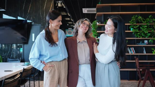 Smiling three multinational businesswomen hugging and looking at camera in office