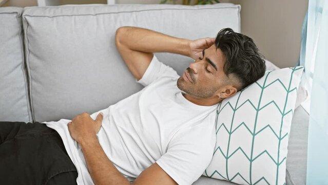 A pained hispanic man in a white shirt lying on a couch indoors clutching his stomach and forehead.