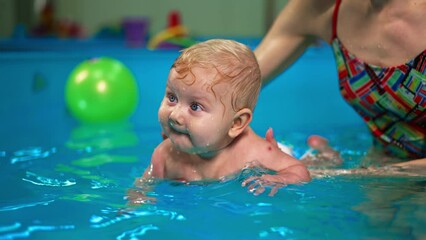 Adorable blond baby boy held by his mom in the water of swimming pool. Mother teaching her child to swim from birth.