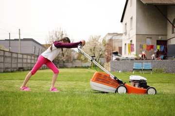 Teenage child girl hardly pushes electric lawnmower during mowing green grass, working in garden on...