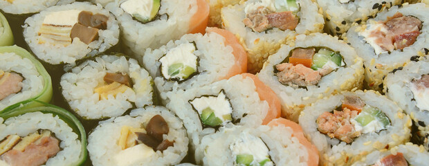 Close-up of a lot of sushi rolls with different fillings lie on a wooden surface. Macro shot of cooked classic Japanese food with a copy space.