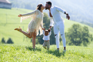 Multiethnic parents and mixed race baby walking on summer grass. Multiethnic couple kissing on...