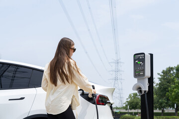 Young woman recharge EV car battery at charging station connected to power grid tower electrical...