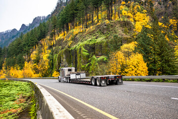 Beige classic big rig semi truck with empty step deck semi trailer driving on the autumn highway...