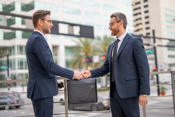 Men in suits handover briefcase outdoor. Business deal. Two business man hold business briefcase. Business transfer deal. Handover of a suitcase in the hands of partners. - obrazy, fototapety, plakaty