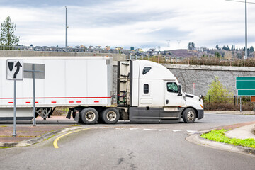 Side view of the white big rig semi truck transporting cargo in dry van semi trailer running on the local road with round intersection