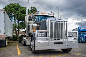 Stylish black powerful big rig semi truck tractor with huge grille guard and empty flat bed semi trailer standing on the truck stop parking lot in row with another semi trucks