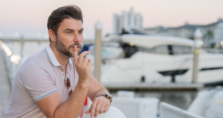 Rich business man dreaming and thinking near the yacht. Well-dressed man wearing fashionable eyeglasses and polo posing outdoor. Male model. Portrait of stylish man in city. Outdoor fashion portrait. - obrazy, fototapety, plakaty