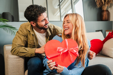 Boyfriend embracing and giving his girlfriend a heart shaped box gift to celebrate Valentines Day. Young couple holding a suprise present on their relationship anniversary. Love and romance date. High