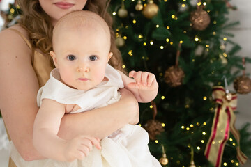 close-up portrait of a little girl in a beautiful white dress in her mother’s arms against the background of a Christmas tree