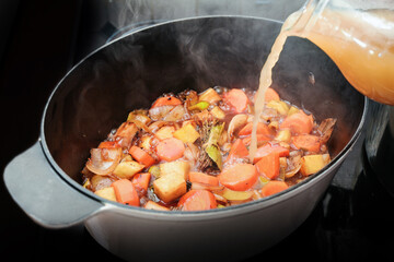 Homemade broth is poured into a casserole pot to deglaze steaming roasted vegetables like carrot,...