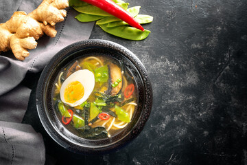 Asia soup with noodles, miso, vegetables, ginger and an egg in a black ceramic bowl on a dark...