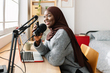 Young african muslim woman recording podcast with laptop and microphone in her room
