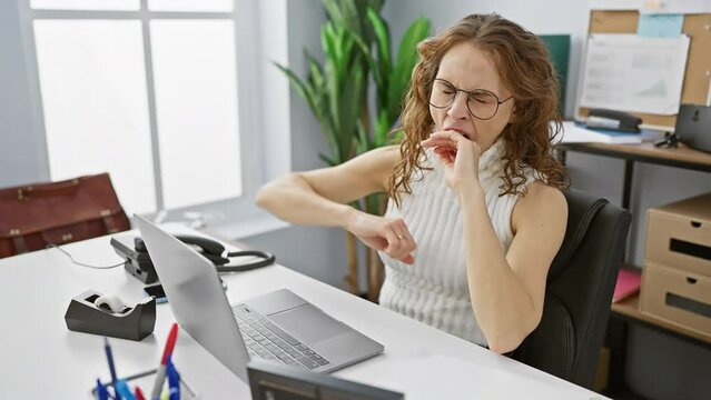 Overworked young woman, bored and yawning at the office, fighting sleepiness with hand over her mouth
