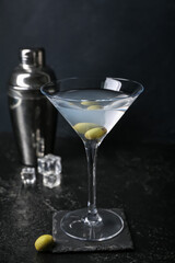 Glass of tasty martini with olives and shaker on dark background