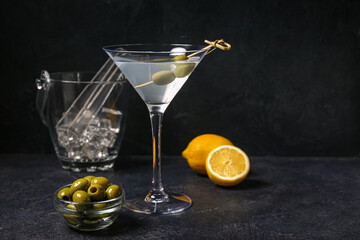 Glass of tasty martini, lemon, ice cubes and green olives on dark background