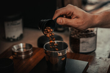 coffee beans being poured into a container, The beans are roasted and have a rich brown color, The scene is set on a wooden surface, and there are blurred objects in the background