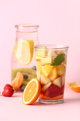 Glass and bottle of infused water with different sliced fruits on pink background
