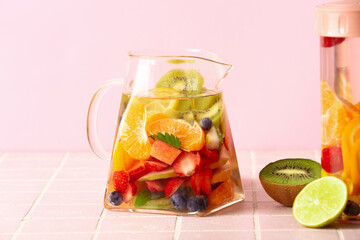 Jug and sports bottle of infused water with different sliced fruits on pink tile table
