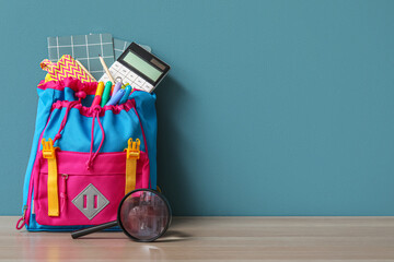 Colorful school backpack with different stationery and magnifier on wooden table near blue wall