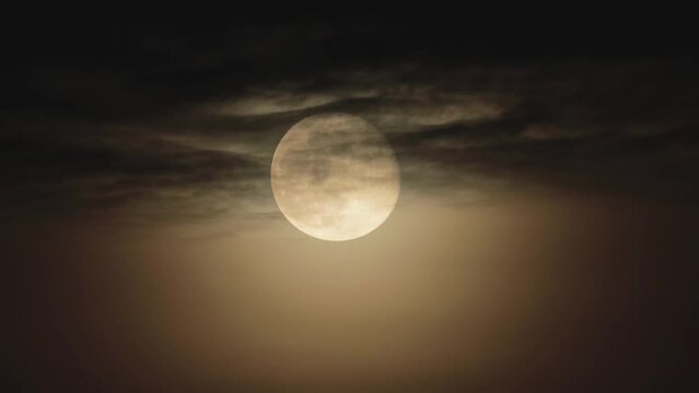 The full moon in the night sky is hidden in dark clouds. The Moon surrounded by an orange glow is diffuse moonlight in the atmosphere