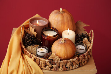 Wicker tray with burning candles, pine cones, pumpkins and autumn leaves on chair near red wall