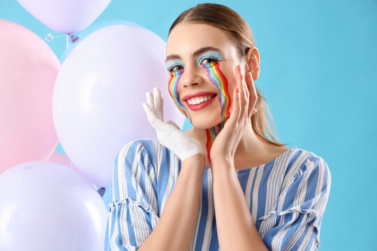 Young woman with painted rainbow on her face and balloons against blue background, closeup