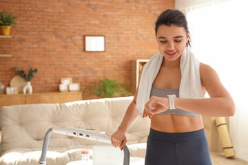 Sporty young woman with treadmill looking at smartwatch in room