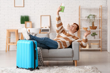 Young man with passport, wooden plane and suitcase lying on sofa at home