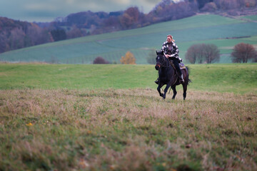 Young woman with red hair gallops with a black horse through an autumnal Young woman with red hair...