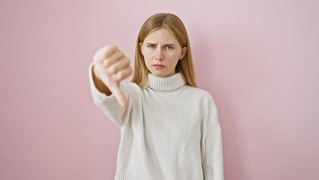 Angry blondie babe in winter sweater shows negative gesture, thumbs down in disgust, face screams 'no!' - perfectly isolated over a pink background.
