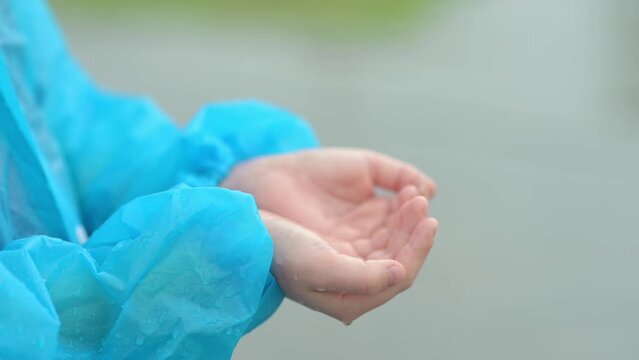Close-up of a child's hands in the pouring rain. A joyful boy in a blue raincoat tries to catch raindrops in his palms while walking on a rainy day. Children love to run and play in rainy weather