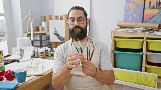 Hispanic man with beard holding paintbrushes in an artist's studio
