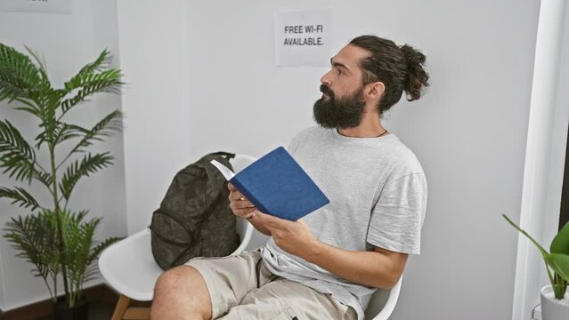 Handsome bearded man reading a book in a modern waiting room with a backpack and plant.